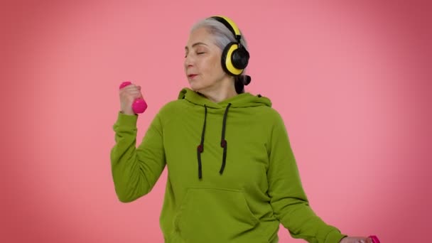 Ανώτερη γιαγιά γυναίκα ακούει μουσική μέσω ακουστικών, γυμνάζεται, σηκώνει ροζ αλτήρες, υγειονομική περίθαλψη — Αρχείο Βίντεο