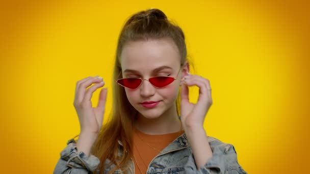 Aufrichtig cooles, fröhliches, stilvolles Mädchen in Jeansjacke mit Sonnenbrille, charmantes Lächeln an der gelben Wand — Stockvideo