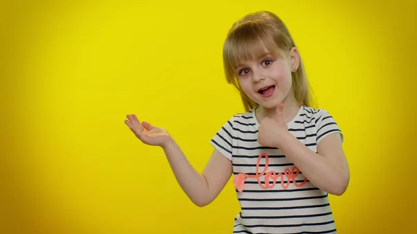 Niña niña mostrando los pulgares hacia arriba y señalando a la izquierda en el espacio en blanco sobre fondo amarillo —  Fotos de Stock