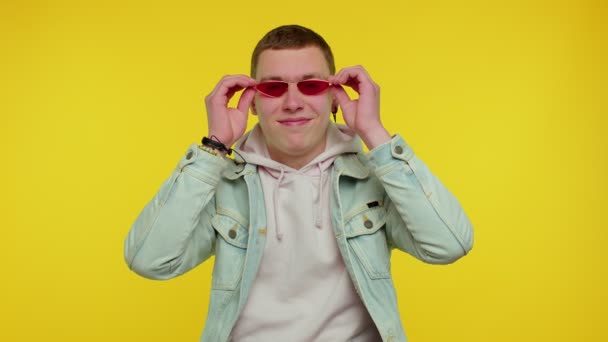 Seductor hombre elegante alegre en chaqueta de mezclilla con gafas de sol, sonrisa encantadora en la pared amarilla — Vídeos de Stock
