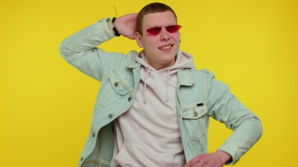 Aufrichtig cooler, fröhlicher, stilvoller Mann in Jeansjacke mit Sonnenbrille, charmantes Lächeln auf gelber Wand — Stockvideo