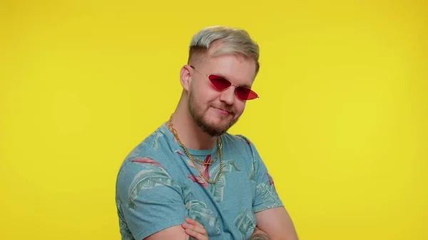 Séduisant homme élégant et joyeux en t-shirt bleu portant des lunettes de soleil, sourire charmant sur le mur jaune — Photo