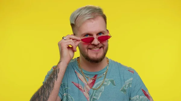 Séduisant homme élégant et joyeux en t-shirt bleu portant des lunettes de soleil, sourire charmant sur le mur jaune — Photo