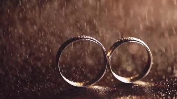 Dos anillos de boda de oro bajo salpicaduras de agua en la superficie oscura que brilla con la luz de cerca macro — Vídeos de Stock