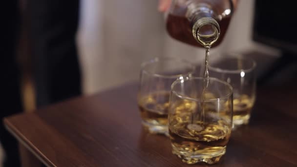 Barman nalewa złotą whisky, koniak, brandy z butelki po alkoholu do szklanek na drewnianym stole — Wideo stockowe