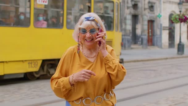 Старшая пожилая туристка в стильной одежде разговаривает по мобильному телефону во время прогулки по городской улице — стоковое видео