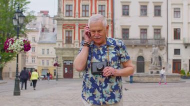 Yaşlı turist şık kıyafetler içinde şehirde yürürken cep telefonuyla konuşuyor.