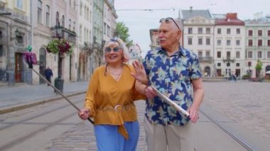 Yaşlı turist ve kadın selfie çubuğuyla şehirde yürüyen ve fotoğraf çeken yaşlı adam.