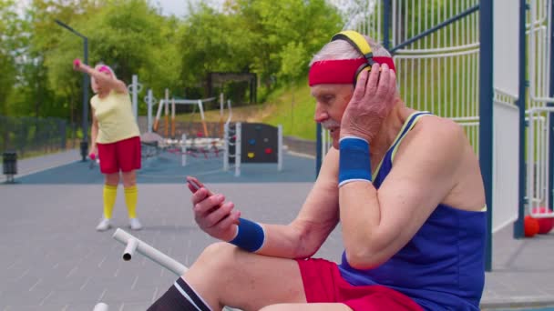 Senior man efter idrottsträning lyssna musik från mobiltelefon bär hörlurar på lekplatsen — Stockvideo