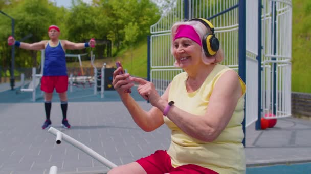 Mulher sênior após treinamento esportivo ouvir música do telefone móvel usando fones de ouvido no playground — Vídeo de Stock