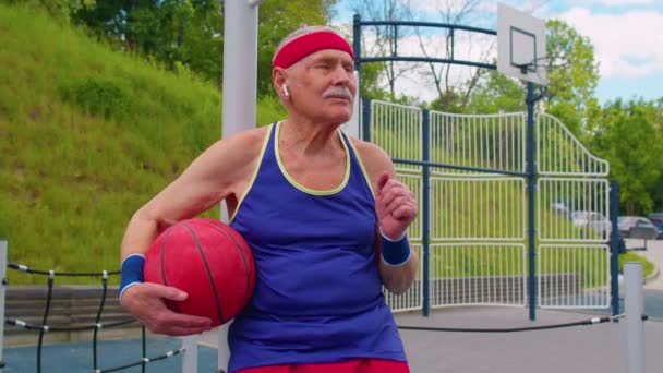 Hombre mayor abuelo después de entrenamiento de baloncesto deportivo sentado, escuchando música en la cancha de juegos — Vídeos de Stock