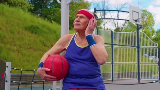 Seniorbestefar etter trening i basketball. Sitte og lytte til musikk på lekeplassen. – stockvideo