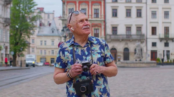 Idősebb férfi turista retro fényképezőgép, mosolygós, zenét hallgatni fülhallgató, tánc az utcán — Stock Fotó