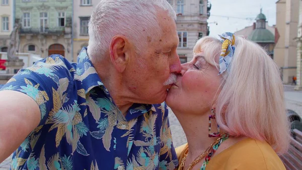 Фотография пары пожилых туристов, делающих селфи в летнем центре города, улыбающихся, глядя в камеру — стоковое фото