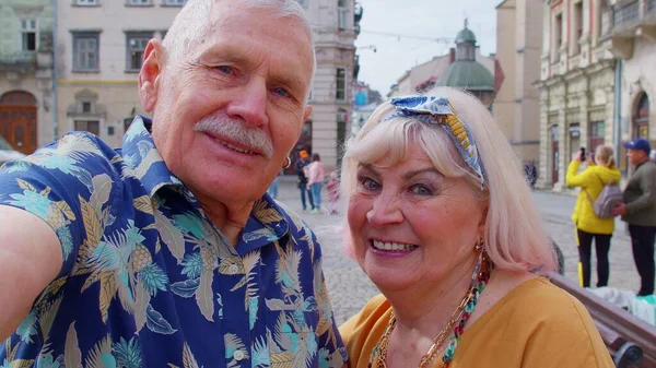 Фотография пары пожилых туристов, делающих селфи в летнем центре города, улыбающихся, глядя в камеру — стоковое фото