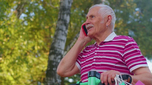 Elderly old grandfather man on electric scooter with colorful shopping bags talking on mobile phone — Stock Photo, Image