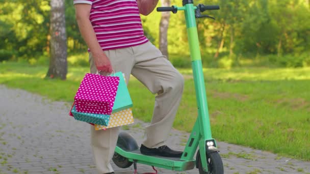 Kakek tua kaukasia bersandar pada skuter listrik setelah berbelanja dengan tas berwarna-warni — Stok Video