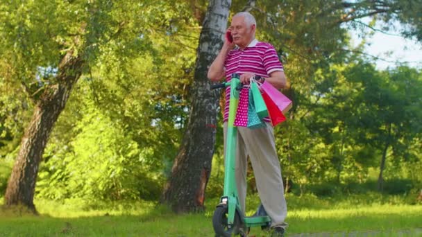 Старший старий дід на електричному скутері з барвистими сумками для покупок, що розмовляють на мобільному телефоні — стокове відео