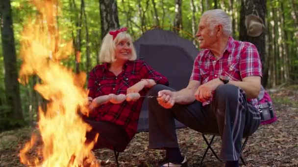 Viejo feliz pareja abuelos descansando en camping en bosque cocinar salchichas sobre fogata — Vídeos de Stock