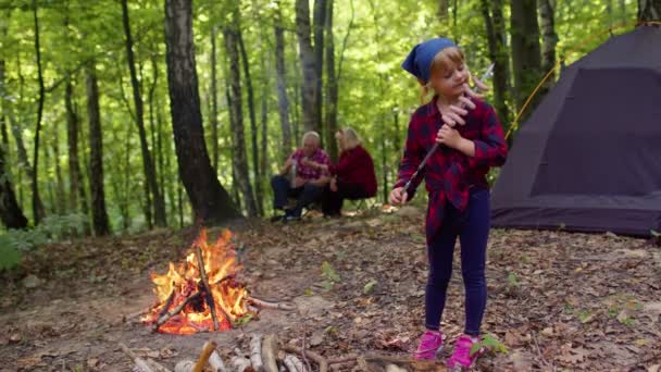 Девочка ест жареные сосиски у костра, дедушка с бабушкой на заднем плане в лесу — стоковое видео