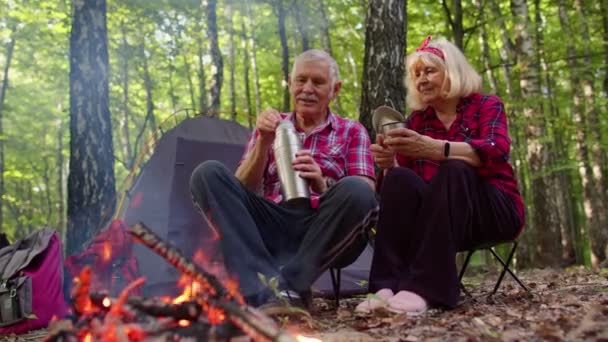 Пожилая бабушка дедушка наливает напиток из термоса, пьет чай над костром в лесу — стоковое видео