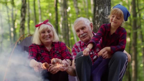 Senior man woman with granddaughter resting at camping in wood cooking frying sausages over campfire — Stock Video