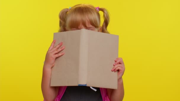 Graciosa niña de la escuela primaria con colas de caballo vistiendo uniforme espiando mientras se esconde detrás de un libro — Vídeos de Stock