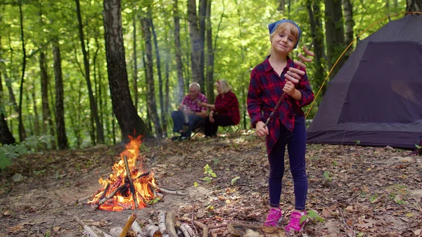 Child girl eating cooked fried sausages near campfire, senior grandparents on background in wood — Stock Photo, Image