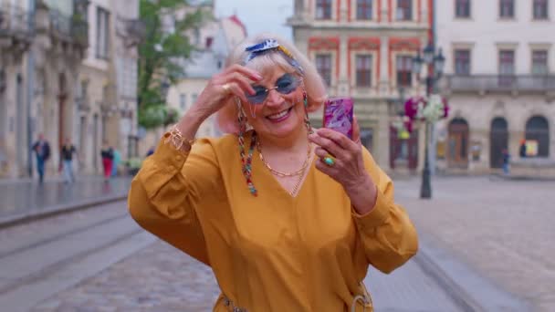 Пожилая бабушка туристка со смартфоном, улыбающаяся, слушающая музыку в приложении в наушниках — стоковое видео