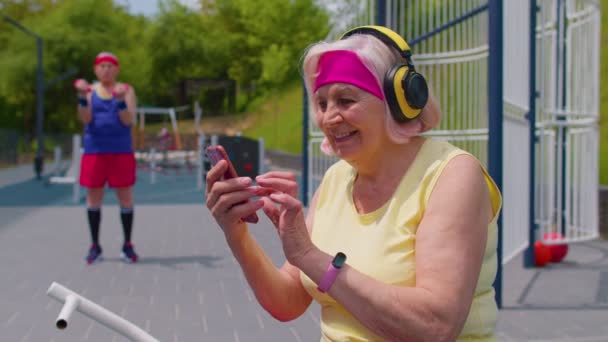 Sénior velho desportista avô com fones de ouvido ouvir música do telefone móvel no playground — Vídeo de Stock