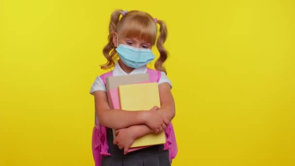 Здорова маленька школярка, яка носить захисну медичну маску, стоїть з книгами, які виглядають втомленими — стокове відео