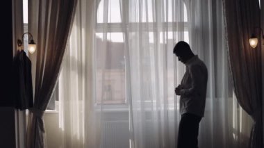 Ceketli adam düğmelerini ilikliyor, beyaz gömleğini ilikliyor dışarı çıkmaya hazırlanıyor, pencerenin yanında yürüyor.