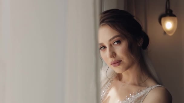 Bride in white dress staying near window and looking at camera, wedding morning preparations at home — Stock Video