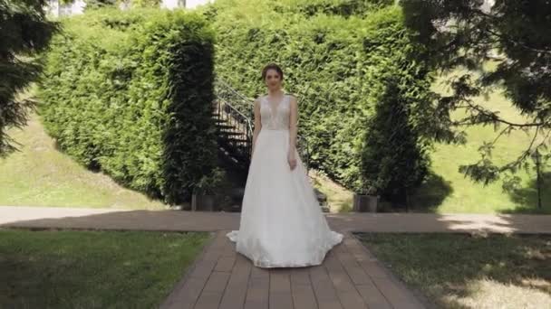 Krásná krásná stylová nevěsta v bílých svatební šaty a závoj procházky v parku a čeká na ženicha — Stock video