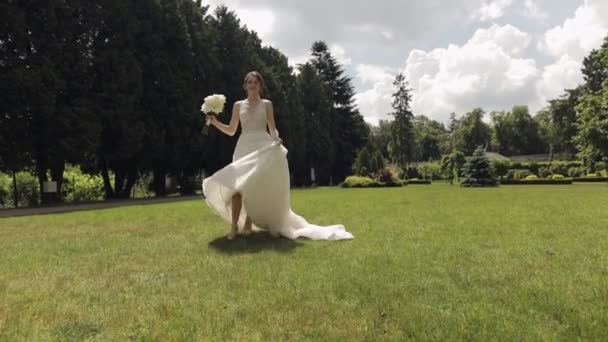 Krásná stylová nevěsta v bílých svatebních šatech a závoj drží svatební kytice v rukou v parku — Stock video