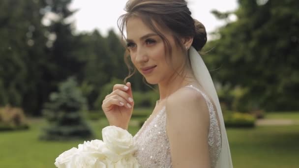 Beautiful stylish bride in white wedding dress and veil holding wedding bouquet in hands in park — Stock Video