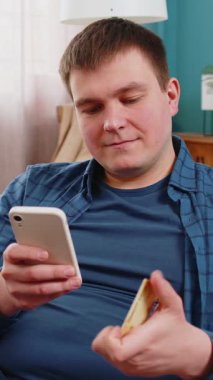 Para transfer ederken kredi kartı ve akıllı telefon kullanan beyaz bir adam, kapalı bir evde çevrimiçi alışveriş nakitsiz yemek siparişi veriyor. Mutlu genç adam kanepede oturuyor..