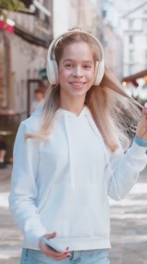 Kablosuz kulaklıkla gezen mutlu genç kadın turistin arka görüntüsü, açık havada akıllı telefondan en sevilen enerjik disko müziğini dinliyor. Şehir sokağının önünden geçen genç bir kızın arka görüntüsü.