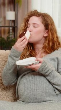 Hamile genç bir kadın, elinde bir parça pasta tutarak kanepesinde oturuyor, neşeli bir ifadeyle yemek yiyor, tatlı tadını çıkarıyor. Tasasız kız küçük zevkleri kucaklıyor, sade mutluluğu.