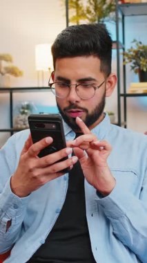 Akıllı telefonu elinde tutan heyecanlı Arap genç adam online bahis oyununun kazanmasıyla hayrete düşmüş iyi haberler okuyor. Kanepede oturan mutlu Hintli adam evdeki galibiyetten çok mutlu bir şekilde ekrana bakıyor..