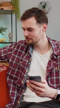 Ekose gömlekli üzgün genç adam elinde akıllı telefon ve kredi kartı kaşları çatık bir şekilde kanepede oturuyor ve internet üzerinden ödeme yapmaya çalışıyor. Parasızlık yüzünden kızgın hissediyor. Kafkasyalı dolandırıcılık kurbanı