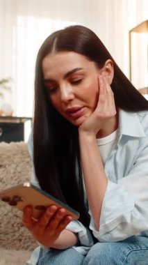 Evde oturan genç bir kadın, üzgün ve hayal kırıklığını gösteren akıllı telefondan mesajlar okuyor. Kanepedeki beyaz kız beklenmedik haberlere duygusal tepki veriyor.