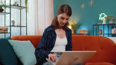 Oturma odasındaki işi bittikten sonra kanepeye oturmuş laptopu kapatan beyaz bir kadın. Mutlu genç kız serbest çalışıyor. Ev kanepesinde uzaktan kumandayla çalışıyor. Defter bilgisayarında e-öğrenme, internette gezinme