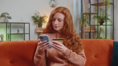 Para transfer ederken kredi kartı ve akıllı telefon kullanan mutlu genç bir kadın. Evin içinde alışveriş yaparken nakit para almadan yemek siparişi veriyor. Kanepede oturan kızıl saçlı kız..