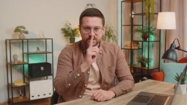 Sessiz olun lütfen. Kafkasyalı genç işadamı işaret parmağını dudaklarına bastırıyor. Ofis masasında sessiz jest işareti yapıyor. Oturma odasında dizüstü bilgisayarlı yetişkin bir adam..