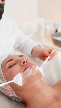 Enzim terapisi. Güzellik uzmanı, genç bir kadının yüzüne fırçayla nemlendirici krem maske takıyor. Kaplıcadaki kız cilt bakımı prosedürü. Yüz kozmetolojisi tedavisi. Dikey video görüntüsü