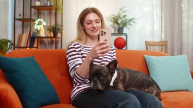 Orta yaşlı bir kadın evde Fransız bulldog 'una sarılıyor ve sosyal medyayı akıllı telefon rahatlama modunda kontrol ediyor. Kanepedeki dişi köpek tomarları besliyor, paylaşıyor, paylaşıyor ve her sabah aynı şeyleri paylaşıyor.