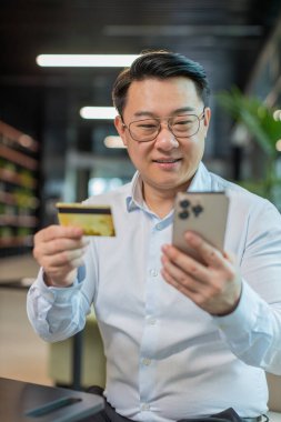 Modern ofis lobisinde online alışveriş ödemeleri için banka kartı ve akıllı telefon kullanan mutlu Çinli işadamı. İş anlaşması. Gülümseyen adam cep telefonu E-bankacılık uygulama servisiyle alışveriş yapıyor..