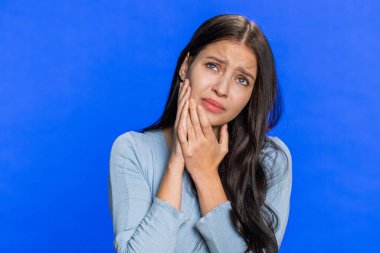 Diş sorunları. Beyaz tenli genç bir kadın yanağa dokunuyor, gözleri korkunç bir ifadeyle kapanıyor diş ağrısı, hassas dişler, çürükler yüzünden acı çekiyor. Güzel kız mavi stüdyo arka planında izole edilmiş.