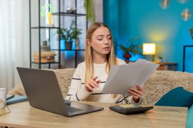 Kadın elinde evraklarla evde oturuyor, hesap makinesi ve dizüstü bilgisayar kullanıyor, vergi beyannamesi üzerinde çalışıyor, istatistiksel veriler tutuyor. Klavyede parmaklarını tıklatan, mali işleri yöneten, istikrar arayan bir kız.